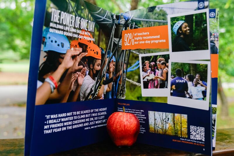 The image shows an open book with a collage of photos related to an outdoor adventure program. There are images of people participating in activities like climbing and team-building exercises. Text excerpts highlight the positive impact of the program, with a focus on overcoming fears and building confidence. A red apple sits in front of the book.